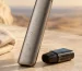 A slim unbranded pod device with water droplets on its casing standing beside its detached pod on a pale stone ledge in warm evening light